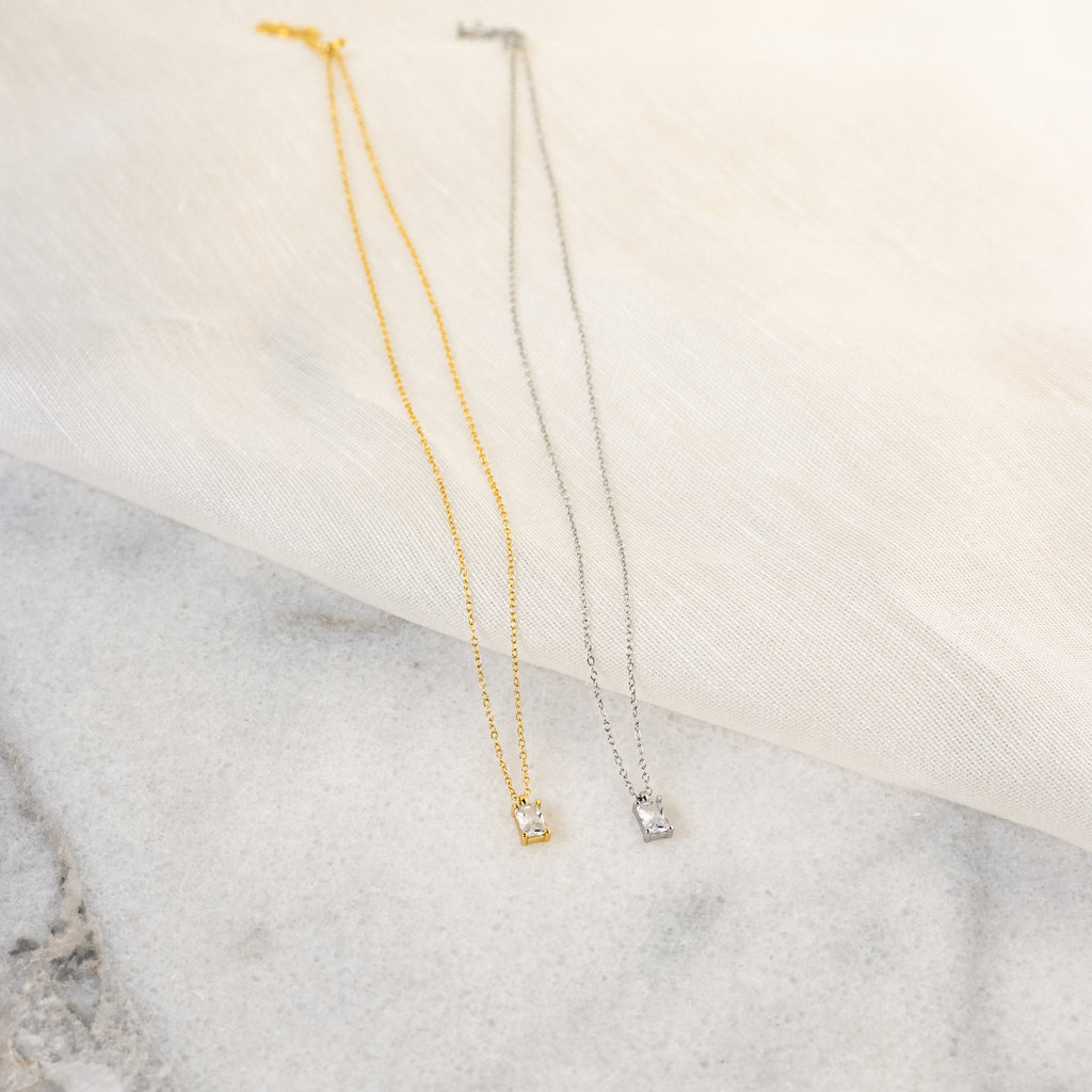 The Sage Diamond Necklace, available in gold and silver with a small square pendant, rests gracefully on fabric and marble—evoking classic diamond necklace elegance.