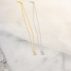 The Sage Diamond Necklace, available in gold and silver with a small square pendant, rests gracefully on fabric and marble—evoking classic diamond necklace elegance.