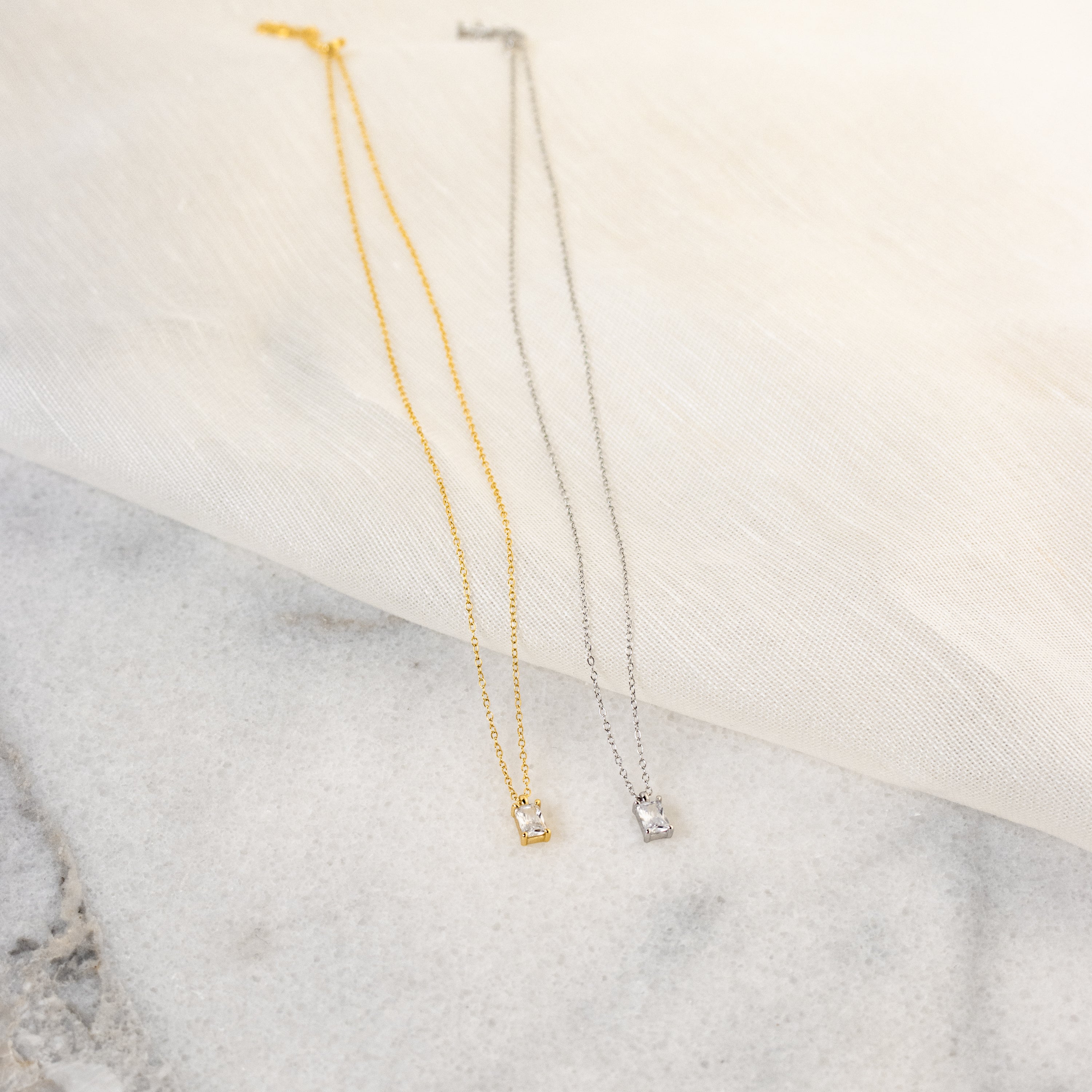 The Sage Diamond Necklace, available in gold and silver with a small square pendant, rests gracefully on fabric and marble—evoking classic diamond necklace elegance.