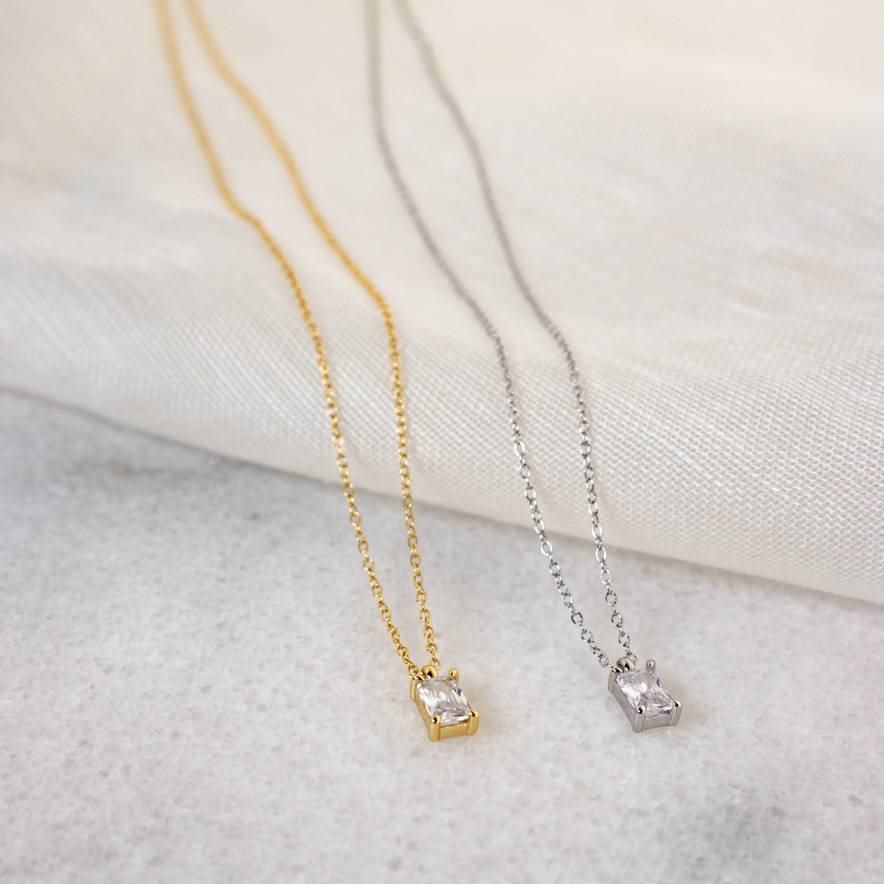 Two Sage Diamond Necklaces—one gold, one silver, each featuring a small rectangular pendant—rest on light fabric, showcasing the refined elegance of this luxury necklace collection.