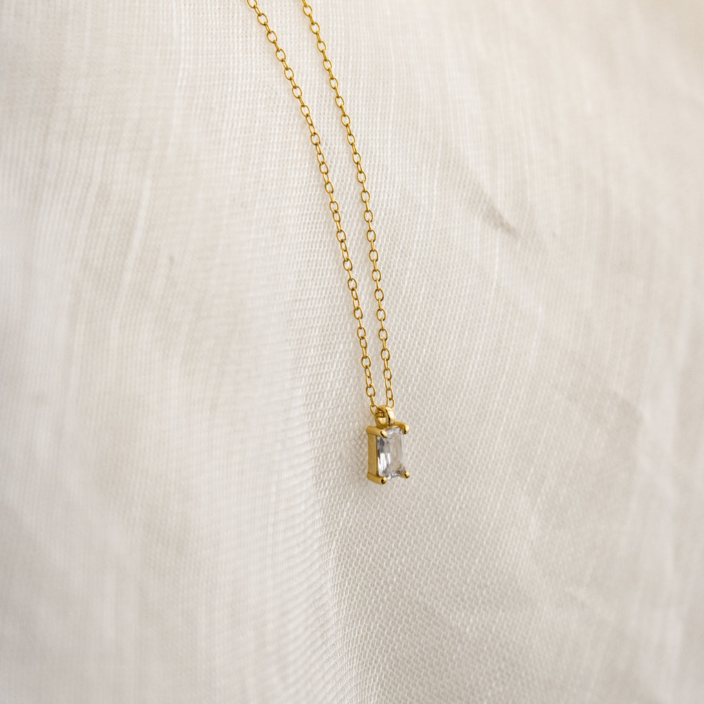 The Sage Diamond Necklace features a delicate gold chain with a rectangular clear gemstone pendant, elegantly displayed against a white fabric background.