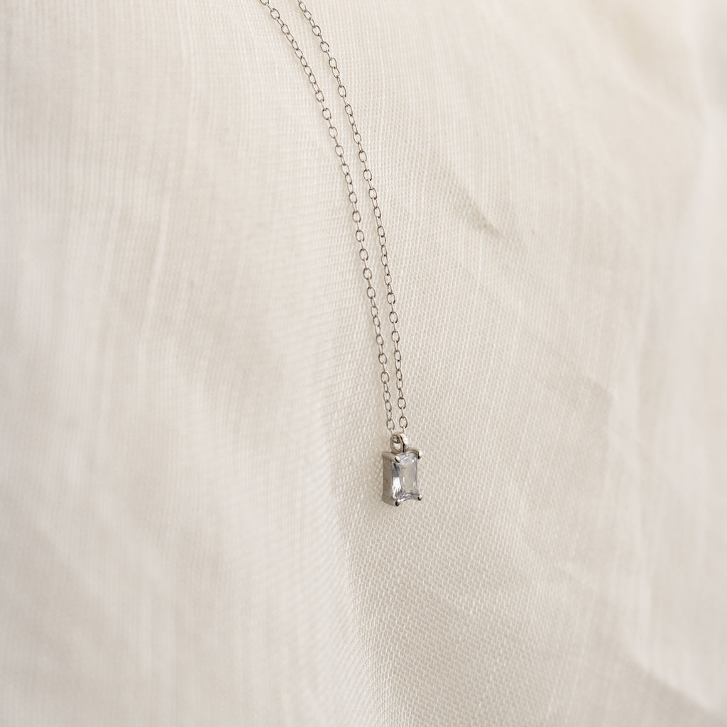The Sage Diamond Necklace in Silver features a delicate chain and rectangular gemstone pendant, radiating timeless elegance against a soft cream fabric background.