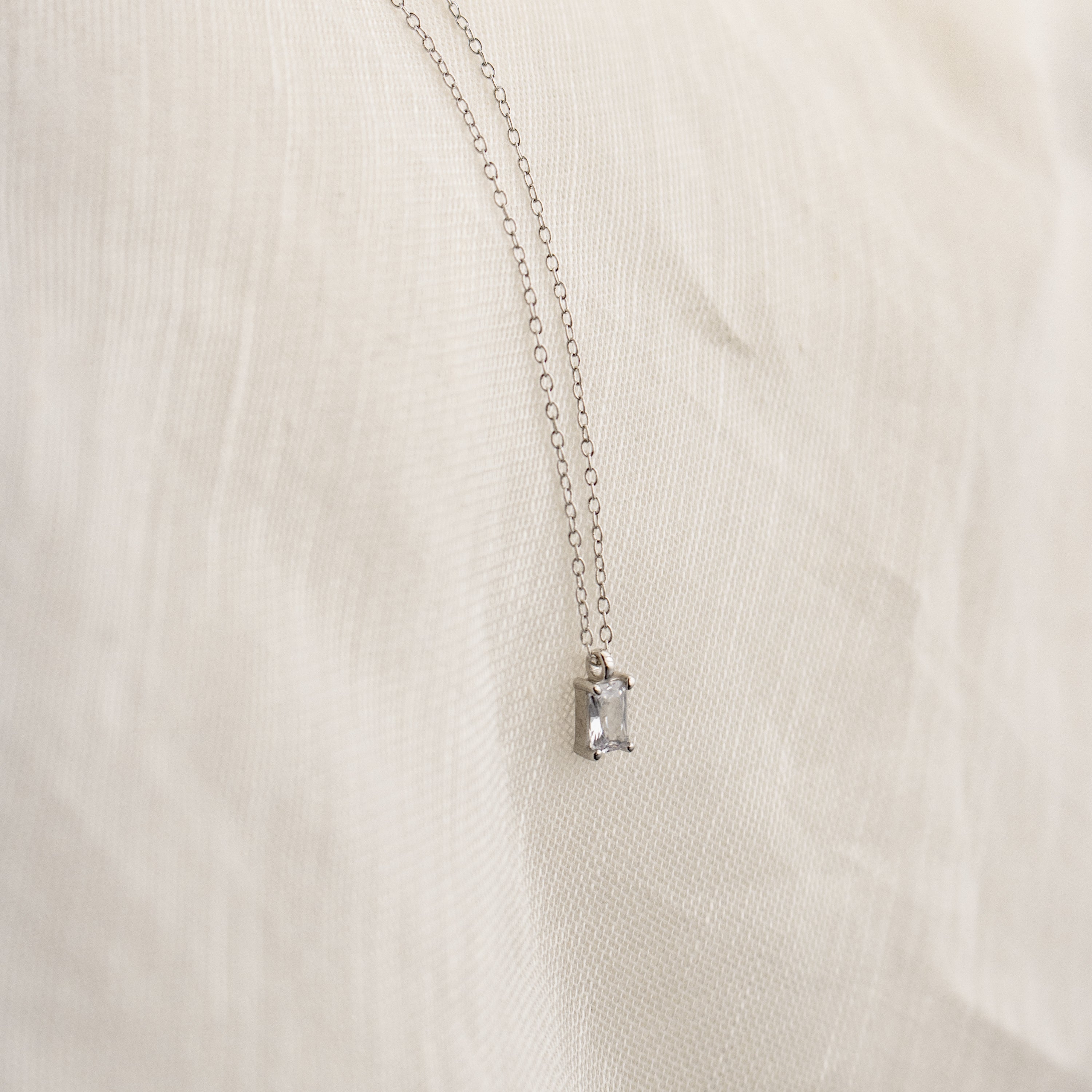 The Sage Diamond Necklace in Silver features a delicate chain and rectangular gemstone pendant, radiating timeless elegance against a soft cream fabric background.