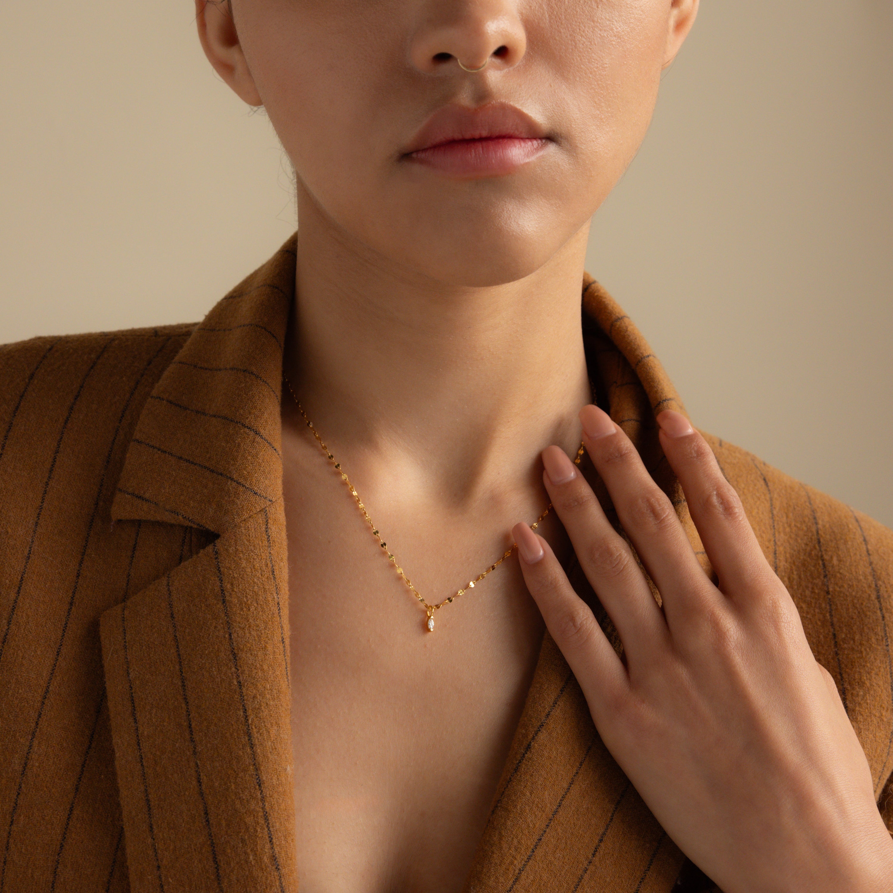 A woman wears a brown pinstripe blazer and the Dainty Marquise Pendant Necklace in 18K Gold, featuring a delicate mirror twist chain, her hand resting near her collarbone.