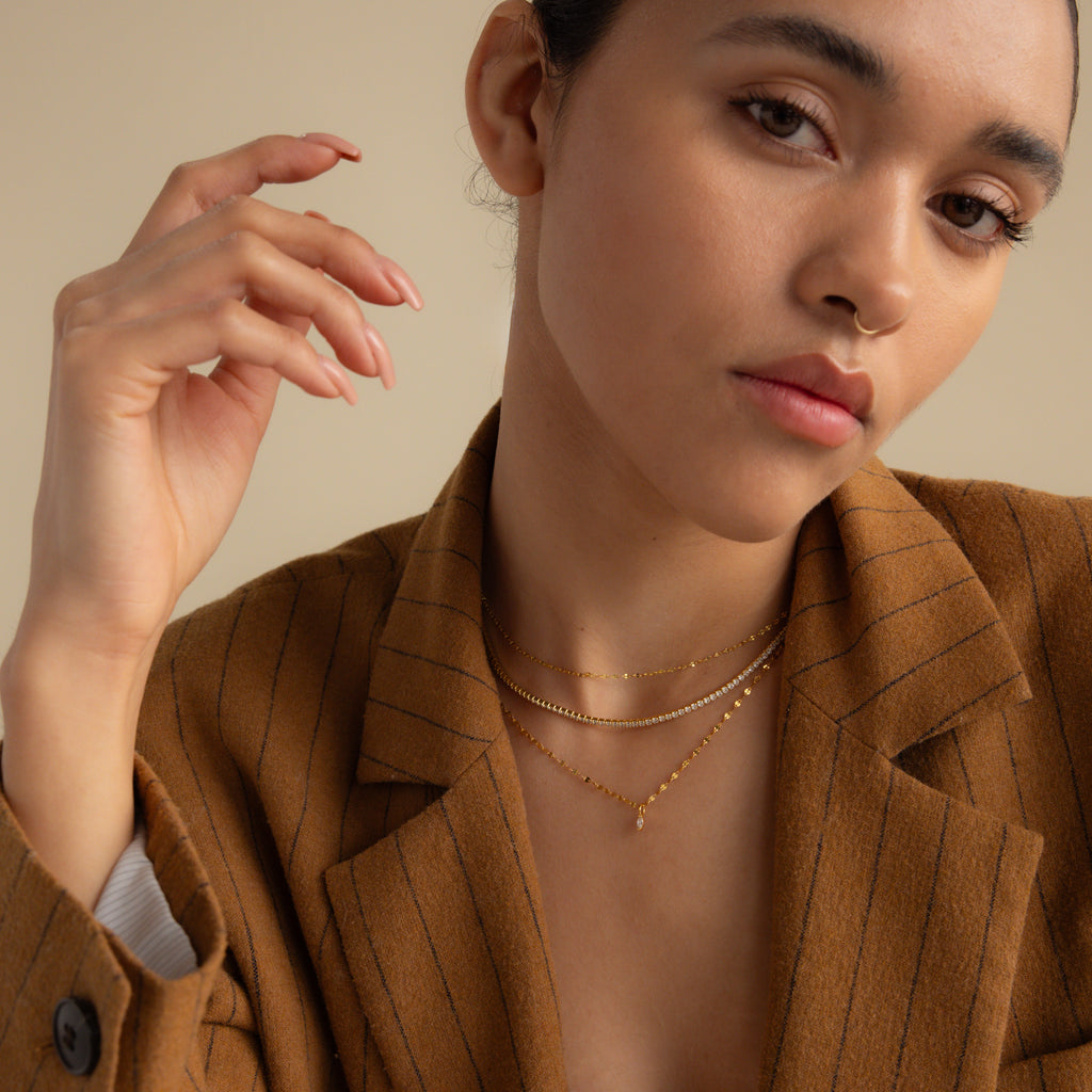 Woman in a brown pinstripe blazer wearing layered gold necklaces, including the Dainty Marquise Pendant Necklace in 18K Gold, posing with her hand raised near her face.
