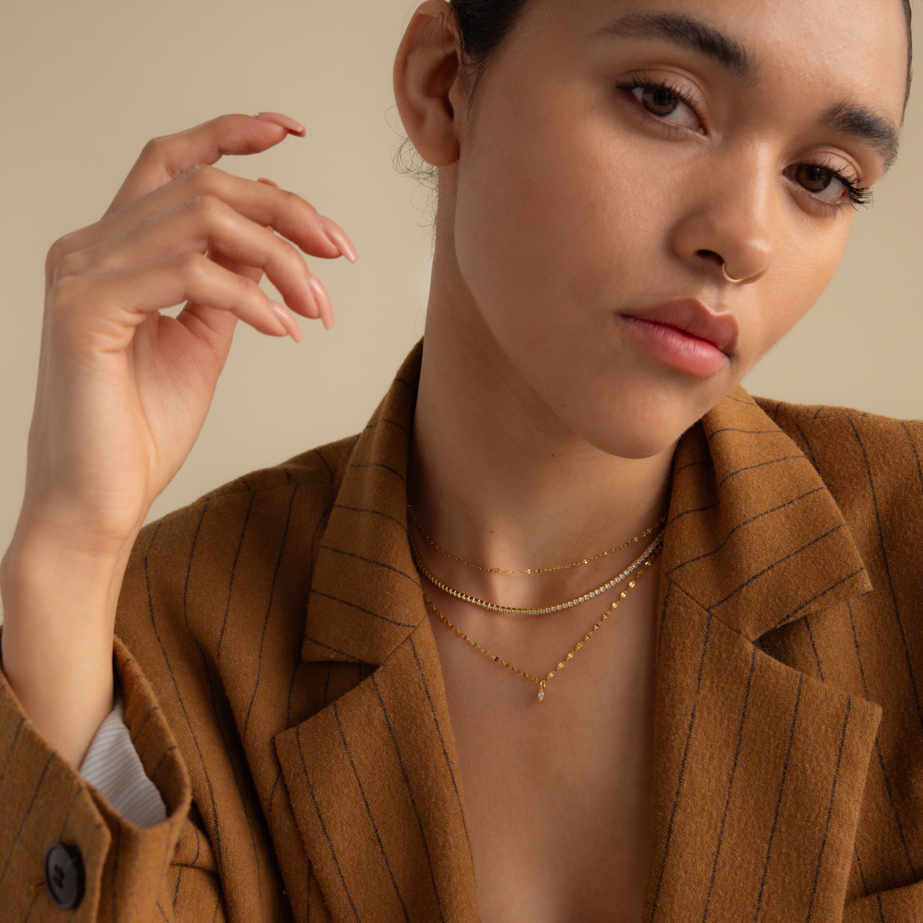 Woman in a brown pinstripe blazer, wearing layered gold necklaces including the Dainty Marquise Pendant Necklace, poses with her hand near her face.