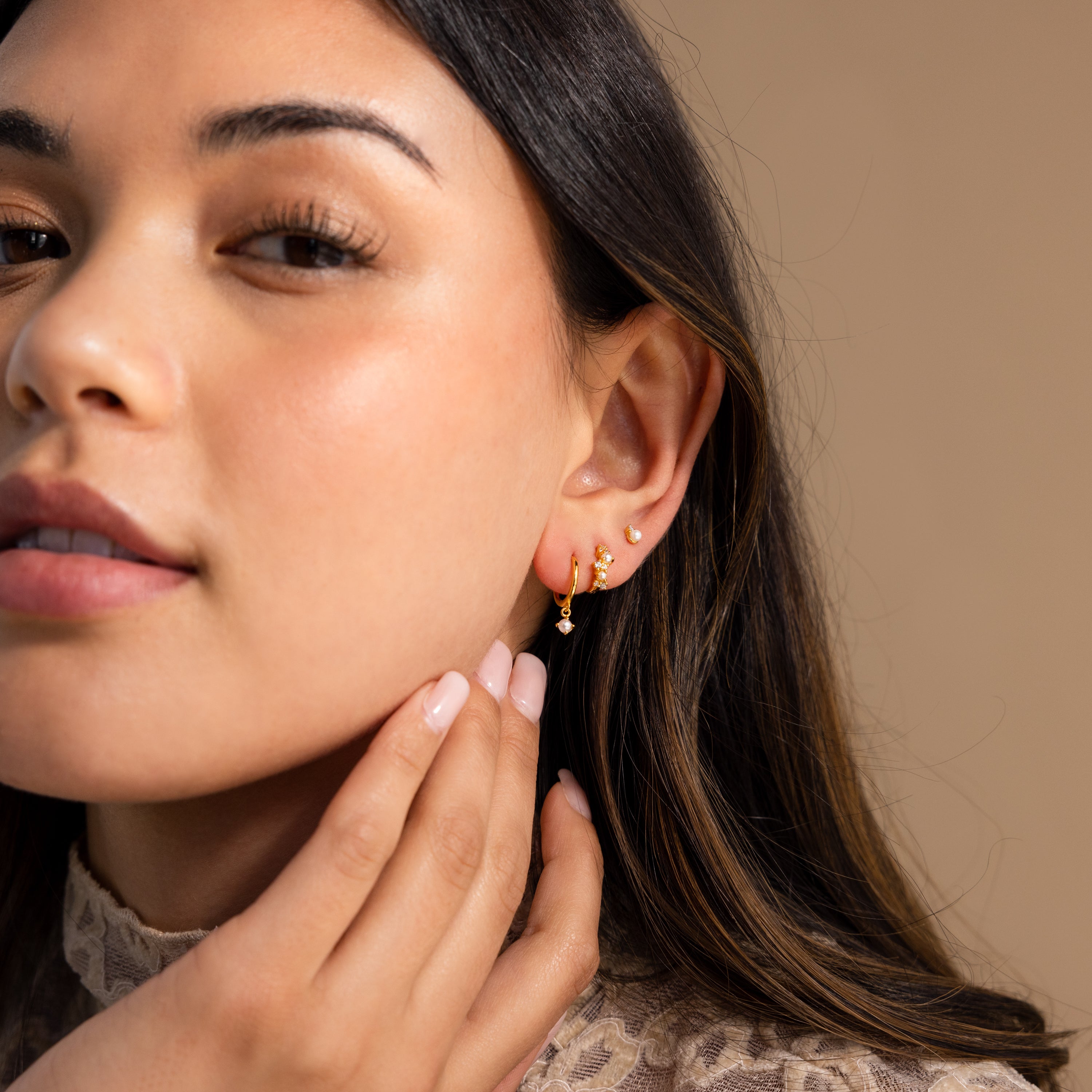 A woman with long dark hair touches her face, wearing Pearl Diamond Cluster Huggies in 18K Gold against a neutral background.