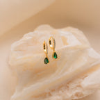 Gold hoop earrings with green birthstone teardrops, shown on a textured white stone background, highlight the elegance of Pave Birthstone Drop Huggies.