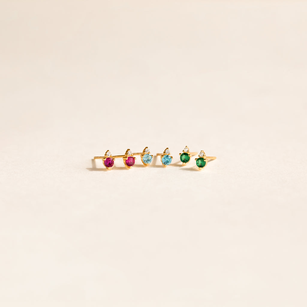 A gold ring with pink, blue, and green gemstones is displayed on a light background next to Raindrop Peridot Studs.