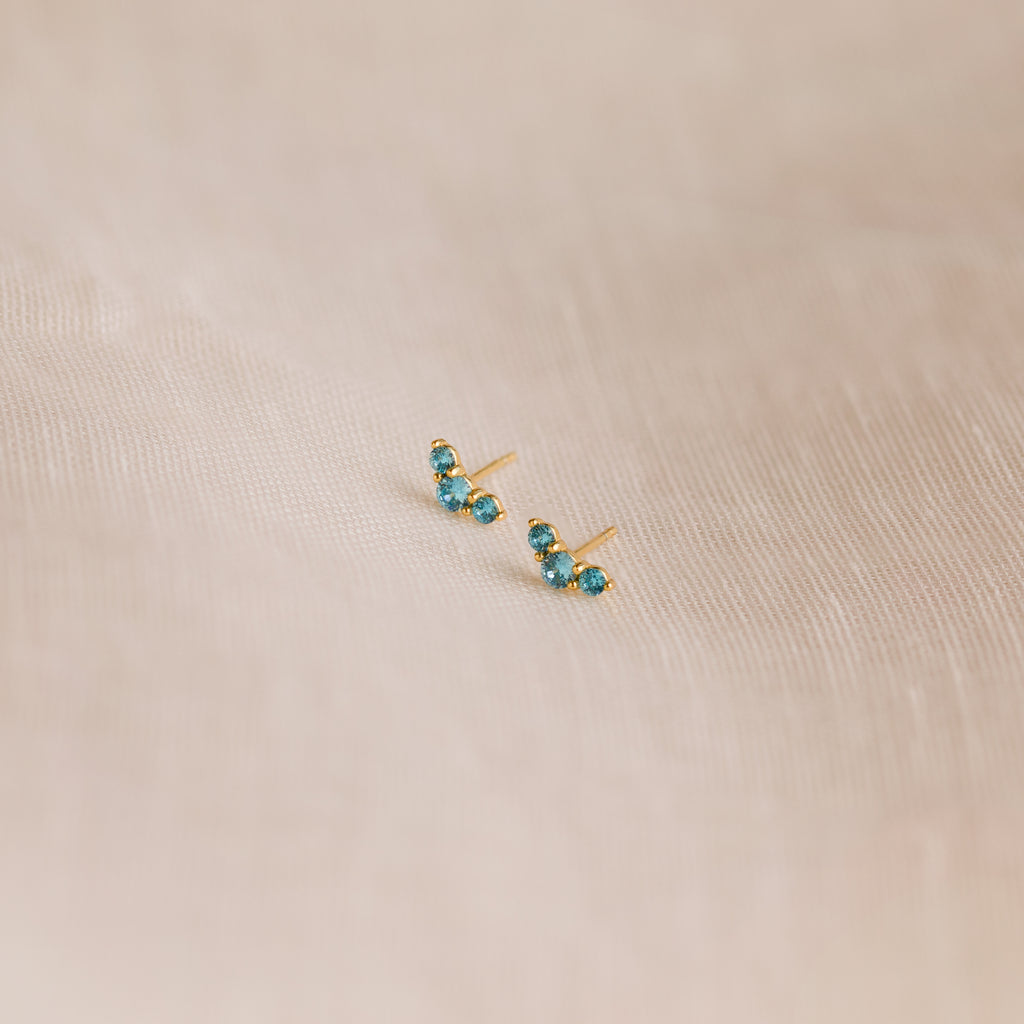 The Bella Blue Zircon Studs feature three sparkling blue gemstones on each gold earring, displayed on soft beige fabric—an ideal gift for mothers and daughters.