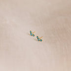 The Bella Blue Zircon Studs feature three sparkling blue gemstones on each gold earring, displayed on soft beige fabric—an ideal gift for mothers and daughters.
