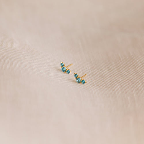 The Bella Blue Zircon Studs feature three sparkling blue gemstones on each gold earring, displayed on soft beige fabric—an ideal gift for mothers and daughters.