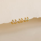 A row of pairs of small gold hoop earrings, each adorned with a square-cut gemstone in 3 colors: light purple, blush pink, and aqua blue, displayed against a textured off-white background.