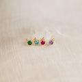 The Raindrop Emerald Studs—dainty gold earrings with green gemstones—are shown on beige textured fabric, perfect for wearing as elegant birthstone studs.