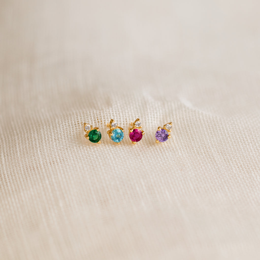 Four Raindrop Peridot Studs with gold settings and green gemstones are arranged in a row on beige fabric.