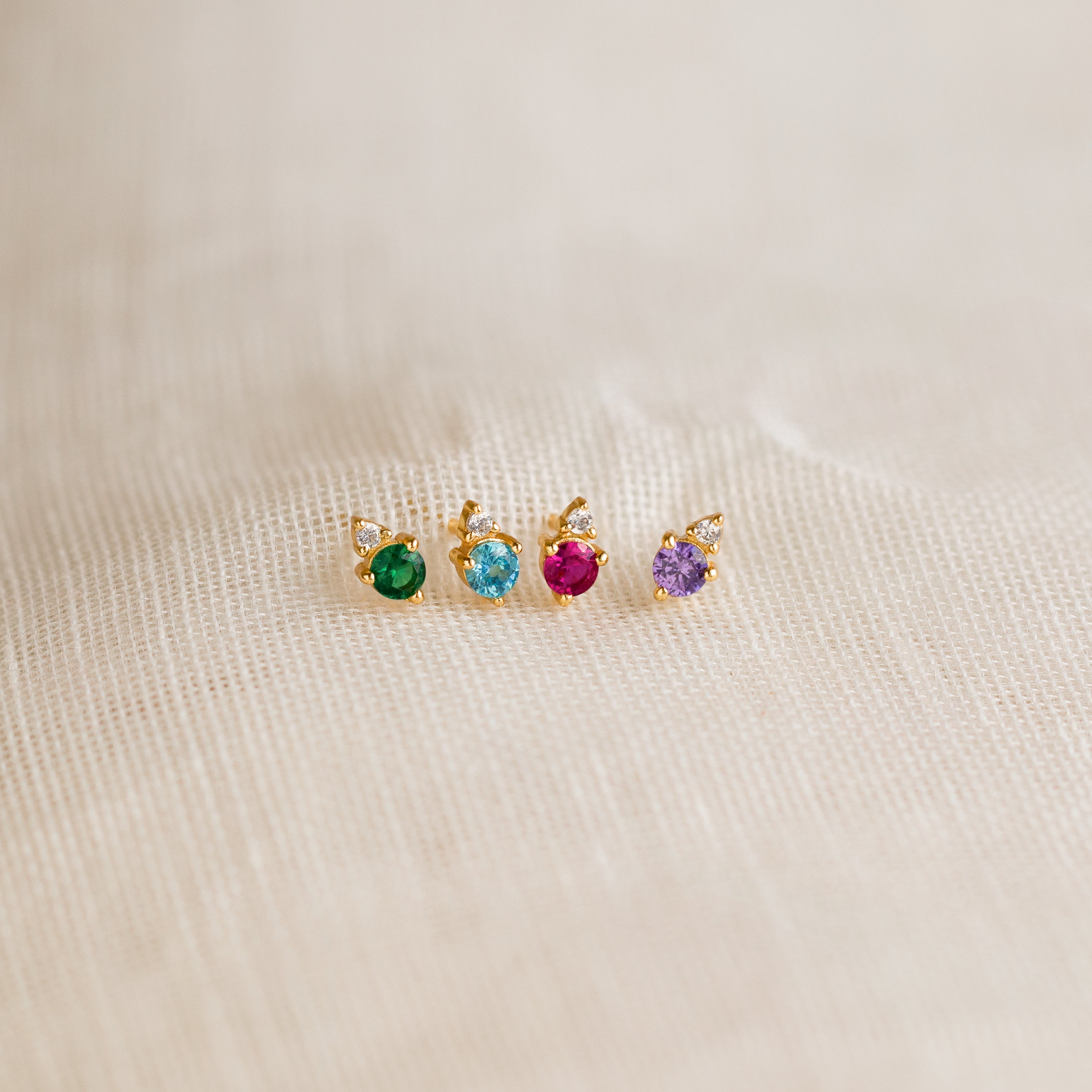 The Raindrop Topaz Studs, featuring green, blue, red, and purple gemstones, are displayed on beige fabric—perfect as colorful birthstone earrings for any occasion.