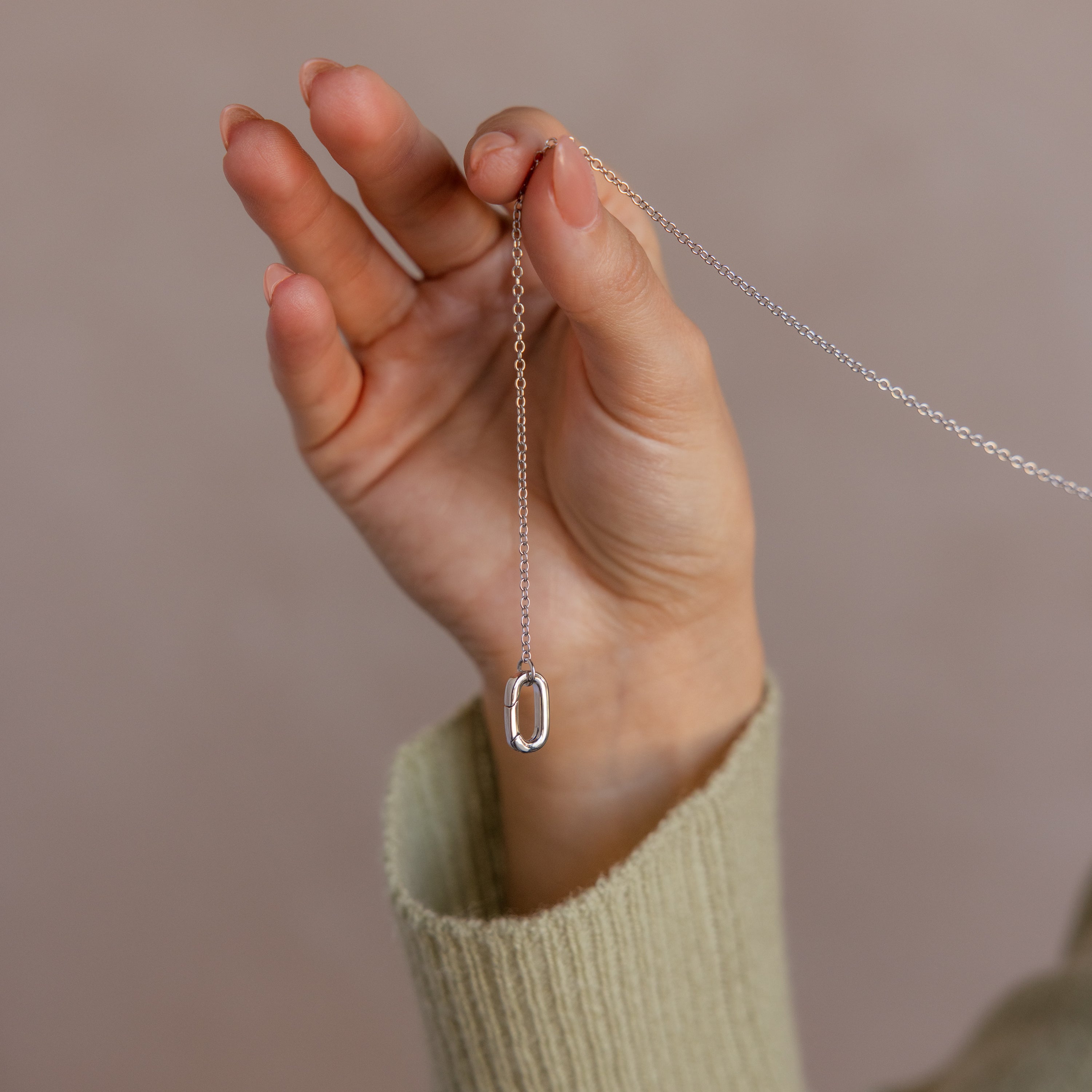 A hand holds the Multipurpose Charm Hoop Clasp Set in White Gold, featuring a delicate oval pendant and charm connector, against a neutral background.