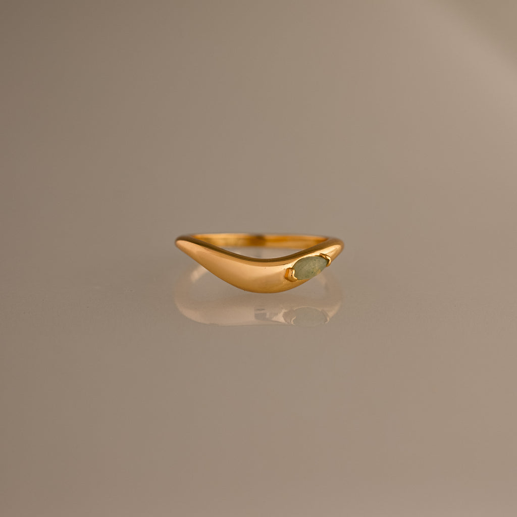 The Zen Jade Curve Ring features a gold sculpted band with a smooth, curved design and a small oval stone set on one side, shown against a neutral background.