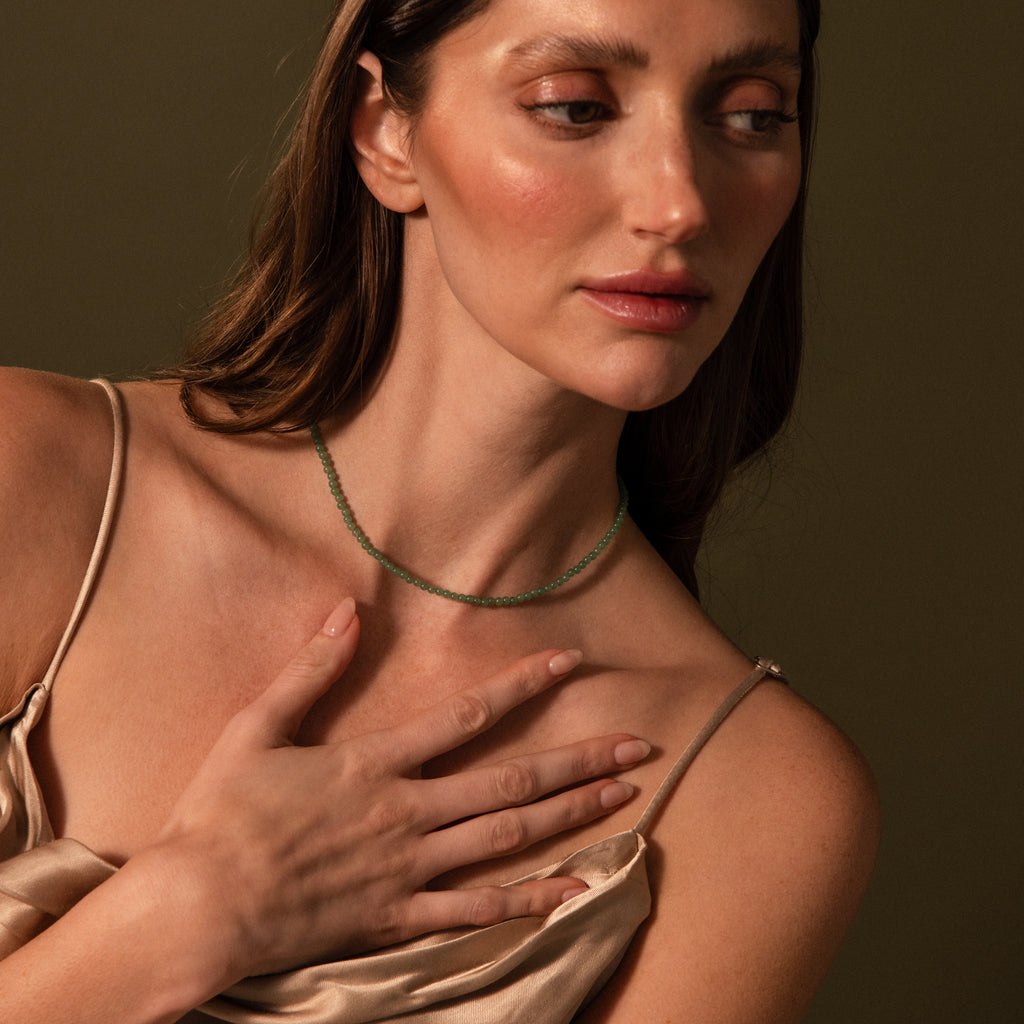 A woman in a satin dress touches her chest, showcasing the Tiny Jade Choker Necklace in 18K Gold against a neutral brown background.
