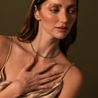 A woman in a satin dress touches her chest, showcasing the Tiny Jade Choker Necklace in 18K Gold against a neutral brown background.
