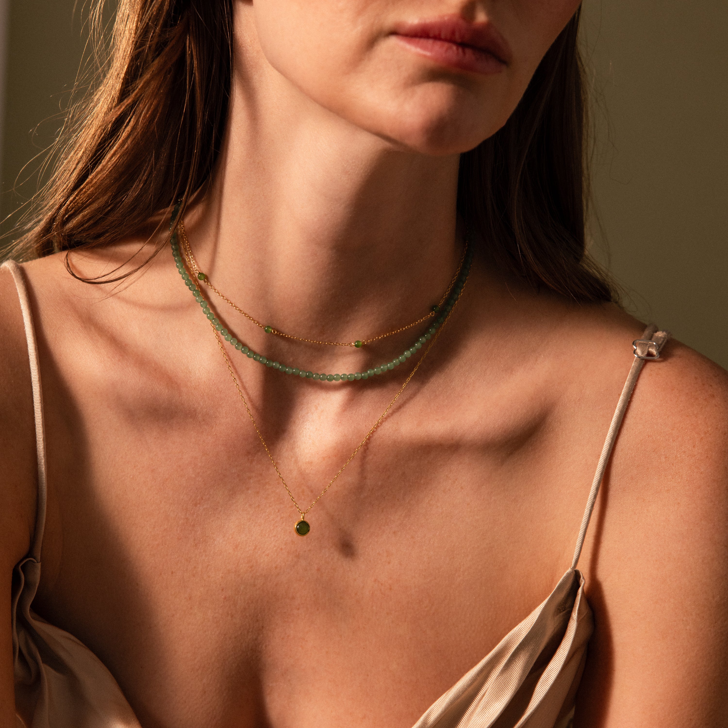 A woman in a beige spaghetti strap top wears layered gold and green necklaces, featuring the Tiny Jade Choker Necklace in Silver, under soft lighting.