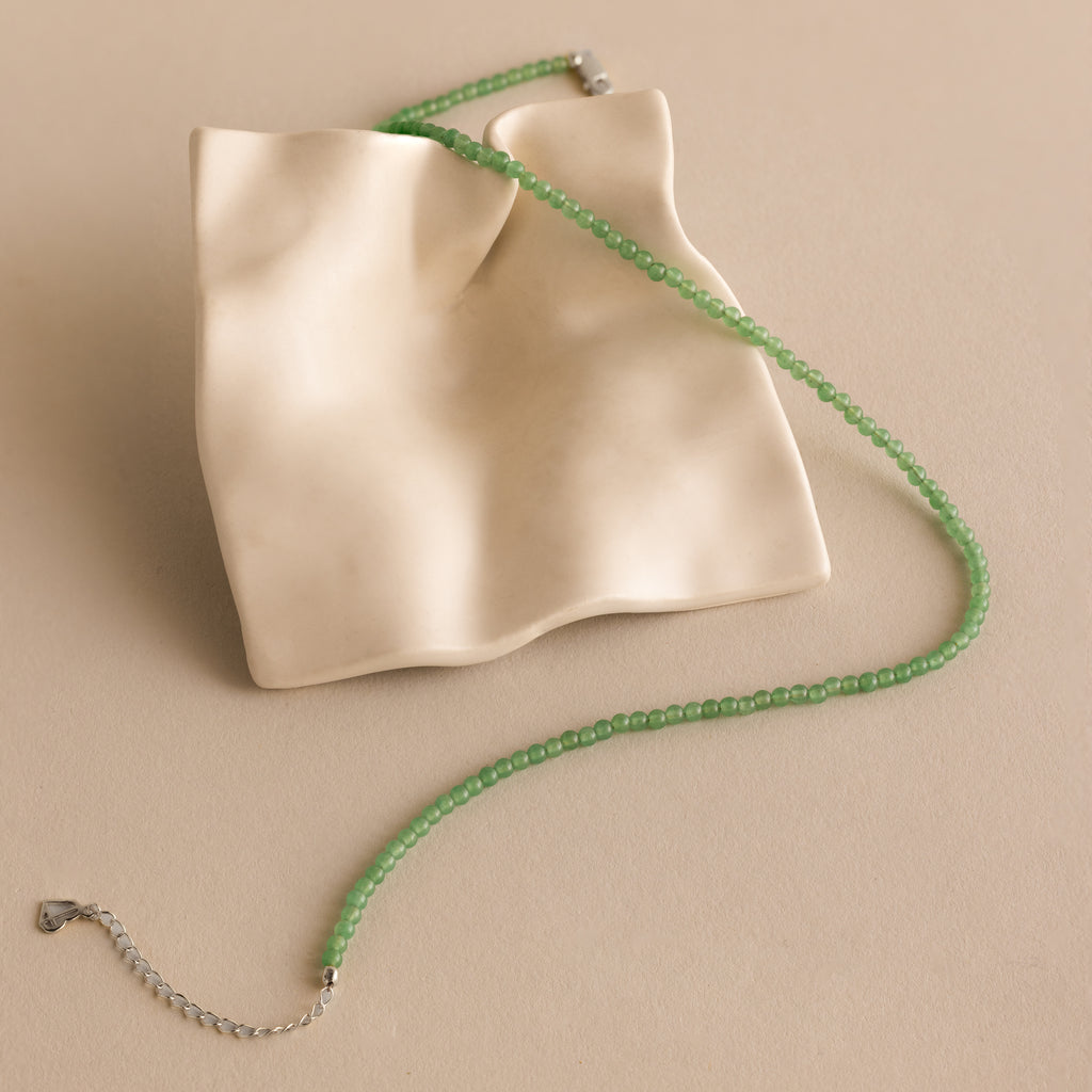 The Tiny Jade Choker Necklace in Silver, featuring green jade beads, is displayed on a draped cream ceramic tray set against a beige background.