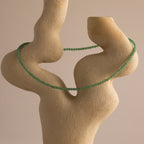 The Tiny Jade Choker Necklace in Silver, featuring green jade beads, is draped over an abstract beige sculpture against a neutral background—ideal for layering elegant necklace looks.