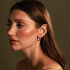 A woman with long brown hair and dewy skin, wearing Noir Pave Dome Hoops in Silver, looks to the left against a muted green background.