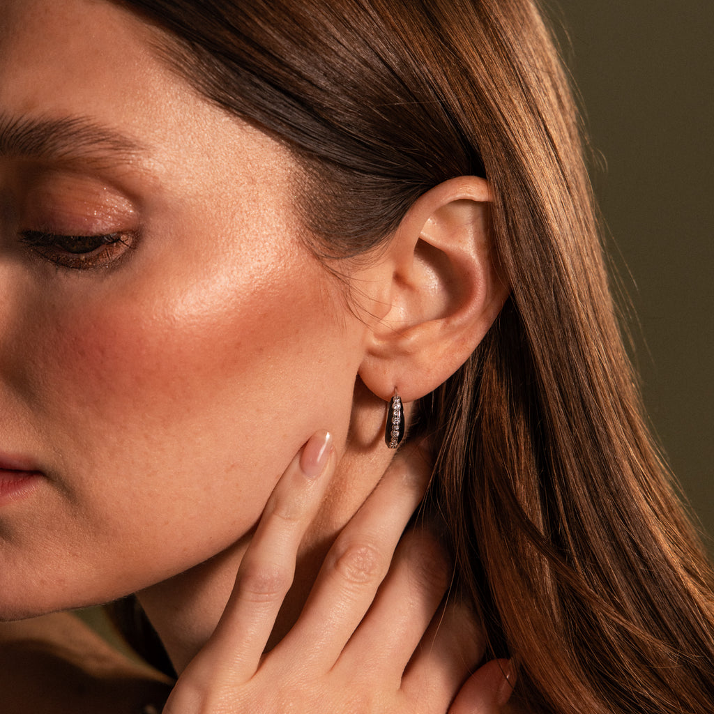 A woman with brown hair touches her face, wearing Noir Pave Dome Hoops—elegant silver earrings adorned with sparkling stones and a refined black enamel accent.