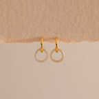 A pair of elegant gold huggie earrings, each featuring a small open circle drop encrusted with sparkling pave stones hang against a minimal, soft beige background with a textured paper edge at the top.