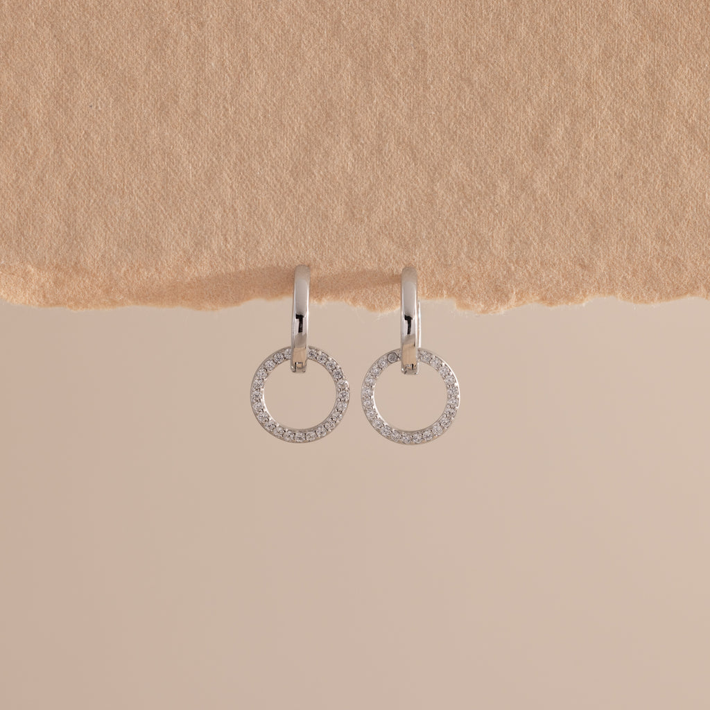 A pair of elegant silver huggie earrings, each featuring a small open circle drop encrusted with sparkling pave stones hang against a minimal, soft beige background with a textured paper edge at the top.