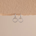 A pair of elegant silver huggie earrings, each featuring a small open circle drop encrusted with sparkling pave stones hang against a minimal, soft beige background with a textured paper edge at the top.