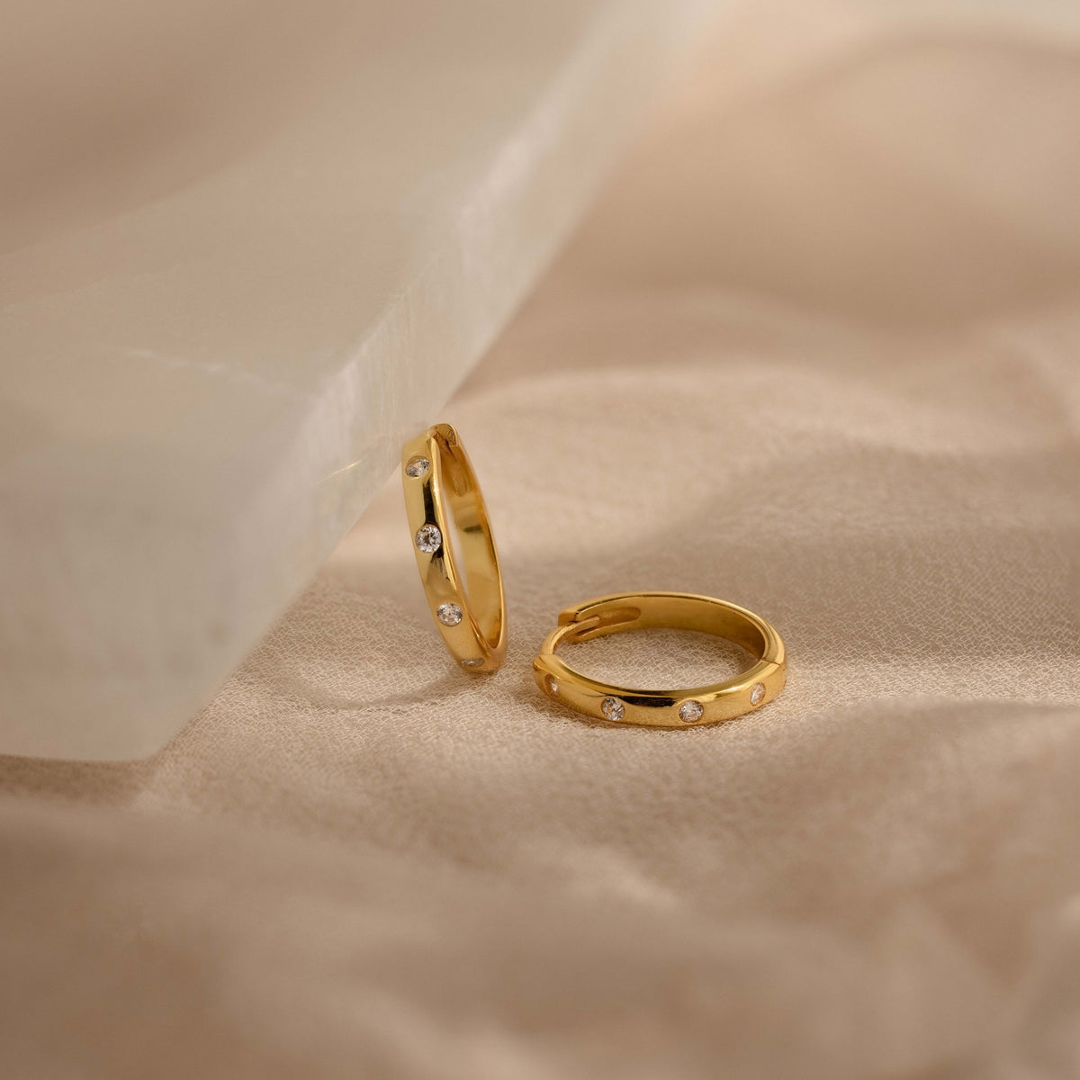 Two gold huggies with small clear stones, displayed on a soft beige fabric surface with a translucent white prop.