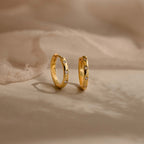 A pair of gold huggie hoop earrings with small clear gemstones set along the front, displayed on a soft beige fabric background.