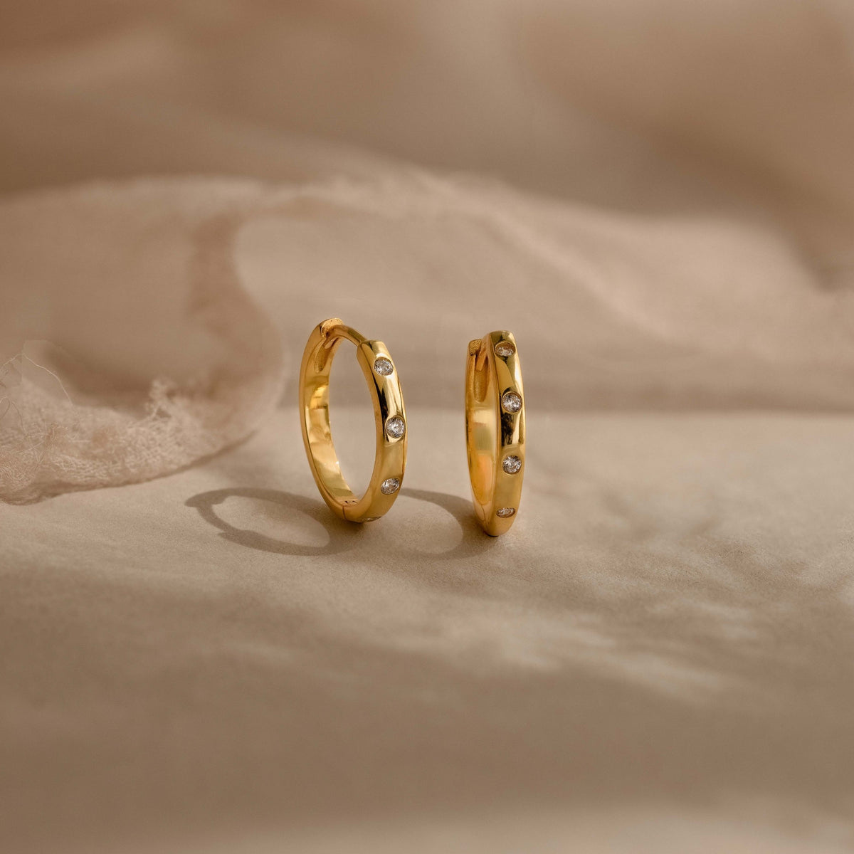 A pair of gold huggie hoop earrings with small clear gemstones set along the front, displayed on a soft beige fabric background.
