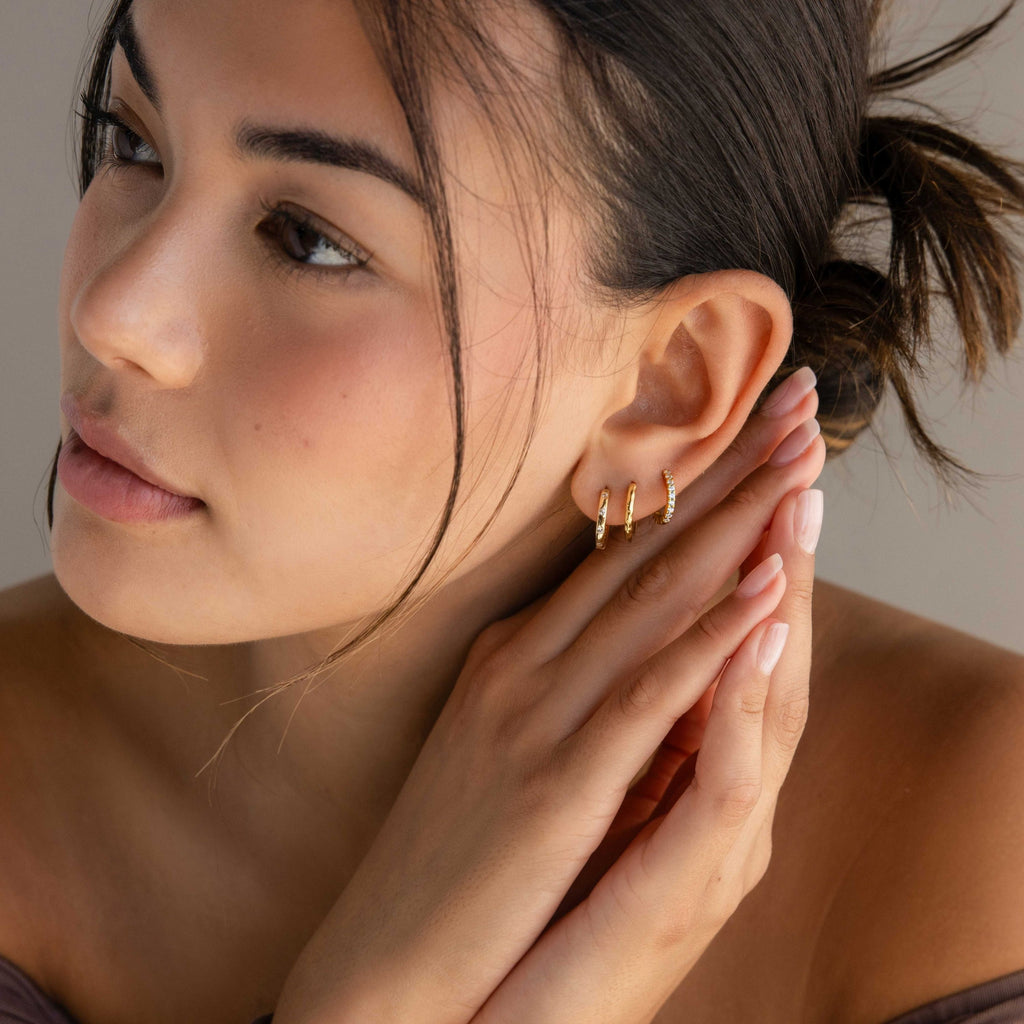 A close-up lifestyle shot of a woman wearing a stack of three gold hoop earrings, including one small gold huggie and one tiny gold pavé diamond stud. She is shown in profile with her hand resting on her neck, wearing a dark brown top against a soft beige background.