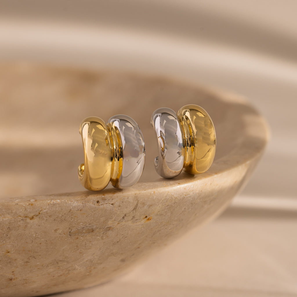 The Mixed Metal Puff Studs, featuring bold gold and silver tones, sit atop a smooth stone surface—perfect for fans of minimal jewelry.