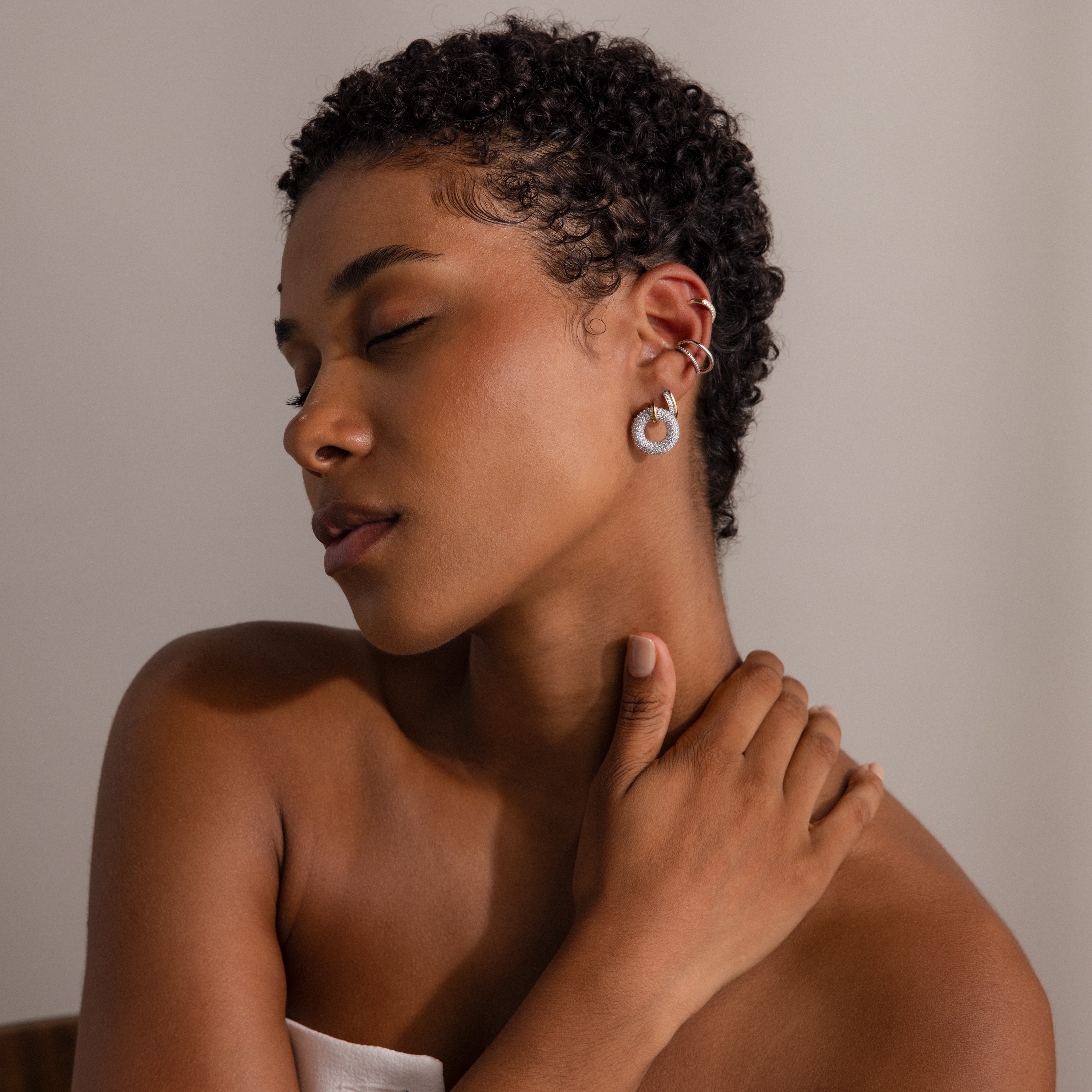 A woman with short curly hair poses with eyes closed, wearing Half Pave Huggies in 18K Gold, gently touching her neck.