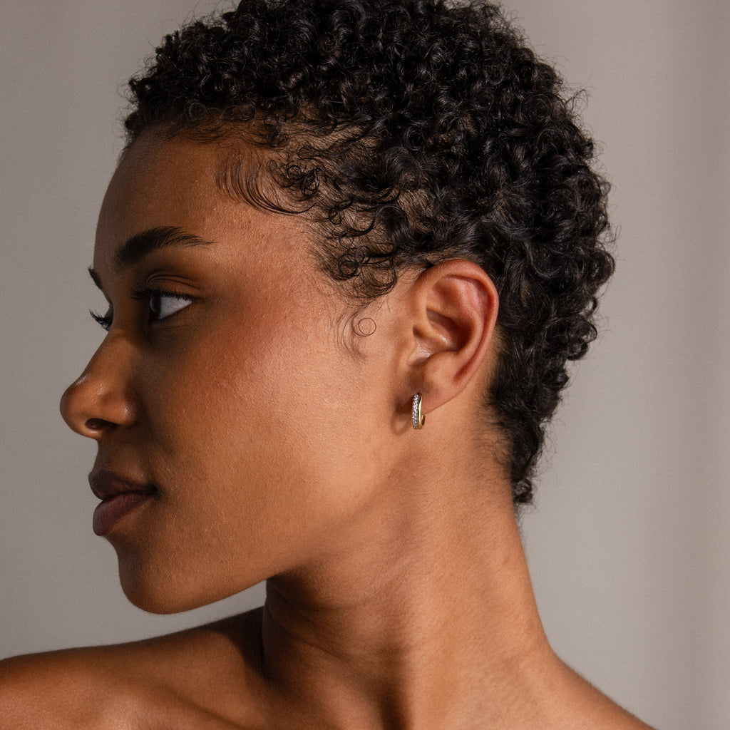 A woman with short curly hair wears shimmering Half Pave Huggies in 18K Gold, gazing to the left against a neutral background.