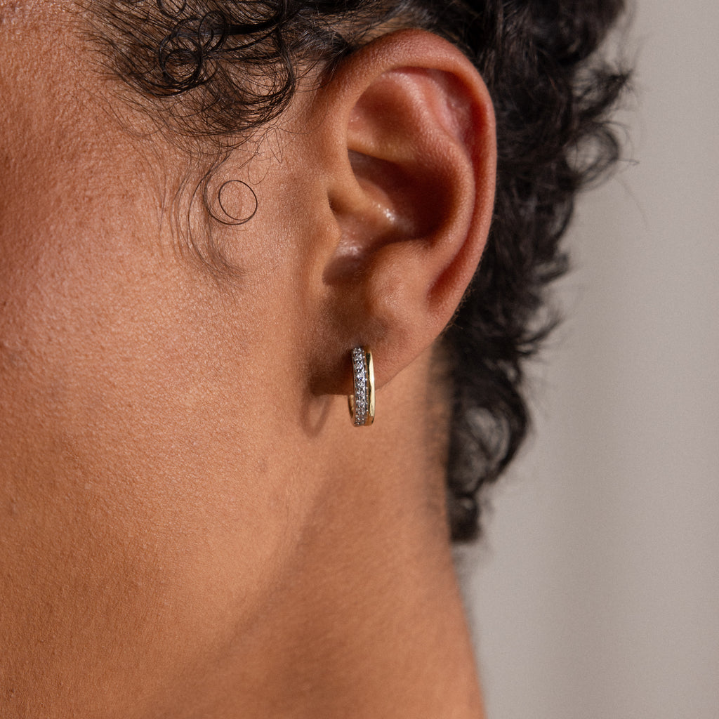 A close-up of a person’s ear wearing a small, textured gold hoop earring, similar to the Half Pave Huggies in 18K Gold.