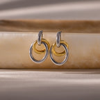 Two pairs of Mixed Metal Linked Earrings, featuring gold and silver tones, rest on a smooth beige stone surface, highlighting their contemporary style ideal for mixing metals.