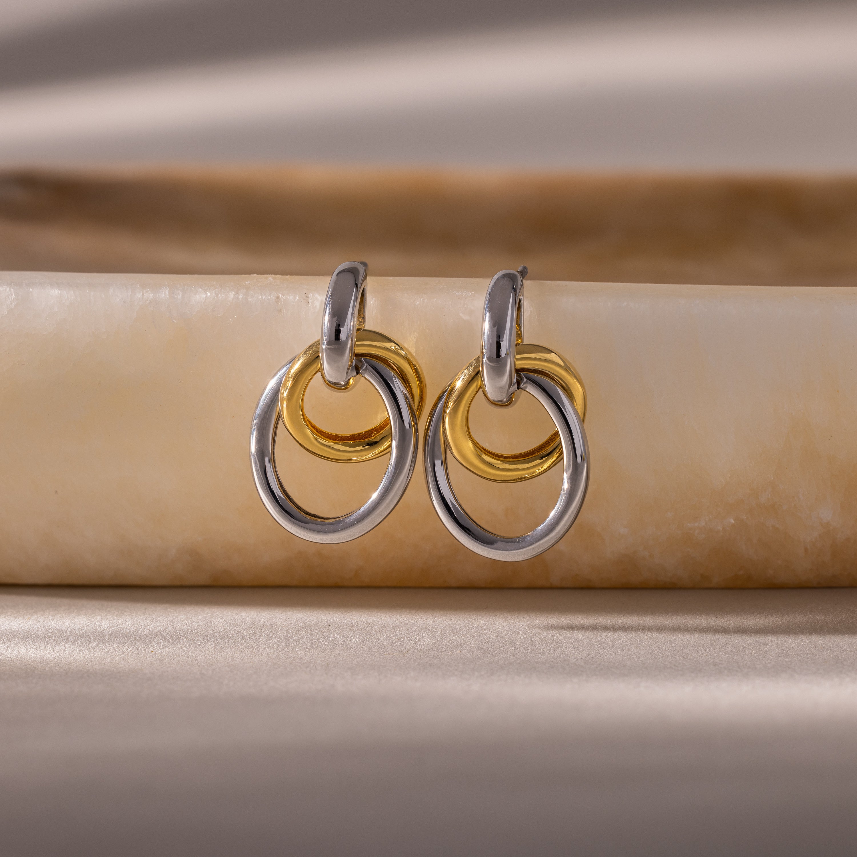 Two pairs of Mixed Metal Linked Earrings, featuring gold and silver tones, rest on a smooth beige stone surface, highlighting their contemporary style ideal for mixing metals.