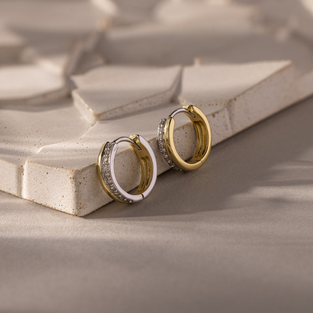 Half Pave Huggies in Mixed Metal, gold and silver earrings with sparkling CZ diamonds, are displayed on a textured, light-colored surface.