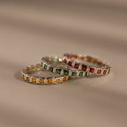 Three Eternity Bezel Birthstone Rings, each with a square cut gemstone in yellow, green, and orange bezel settings, are stacked elegantly on a neutral surface.