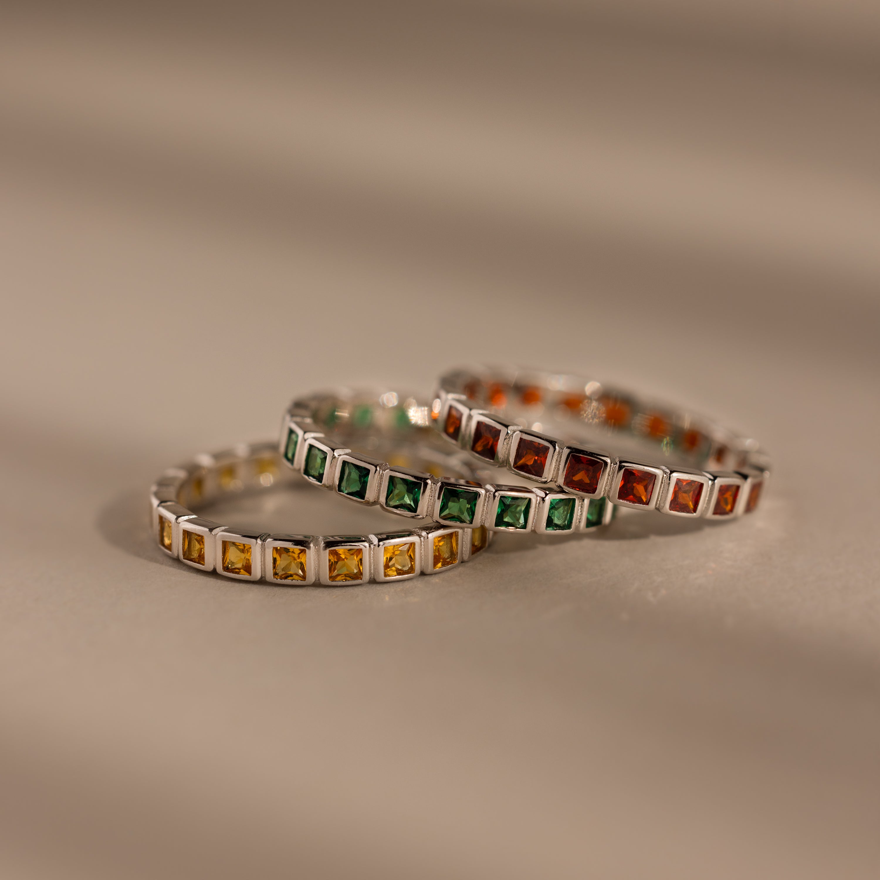 Three Eternity Bezel Birthstone Rings, each with a square cut gemstone in yellow, green, and orange bezel settings, are stacked elegantly on a neutral surface.