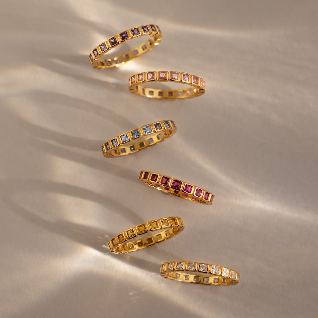 Six Eternity Bezel Peridot Rings with colorful square gemstones are arranged diagonally on a beige surface with soft shadows, highlighting the timeless elegance of this bezel birthstone ring.