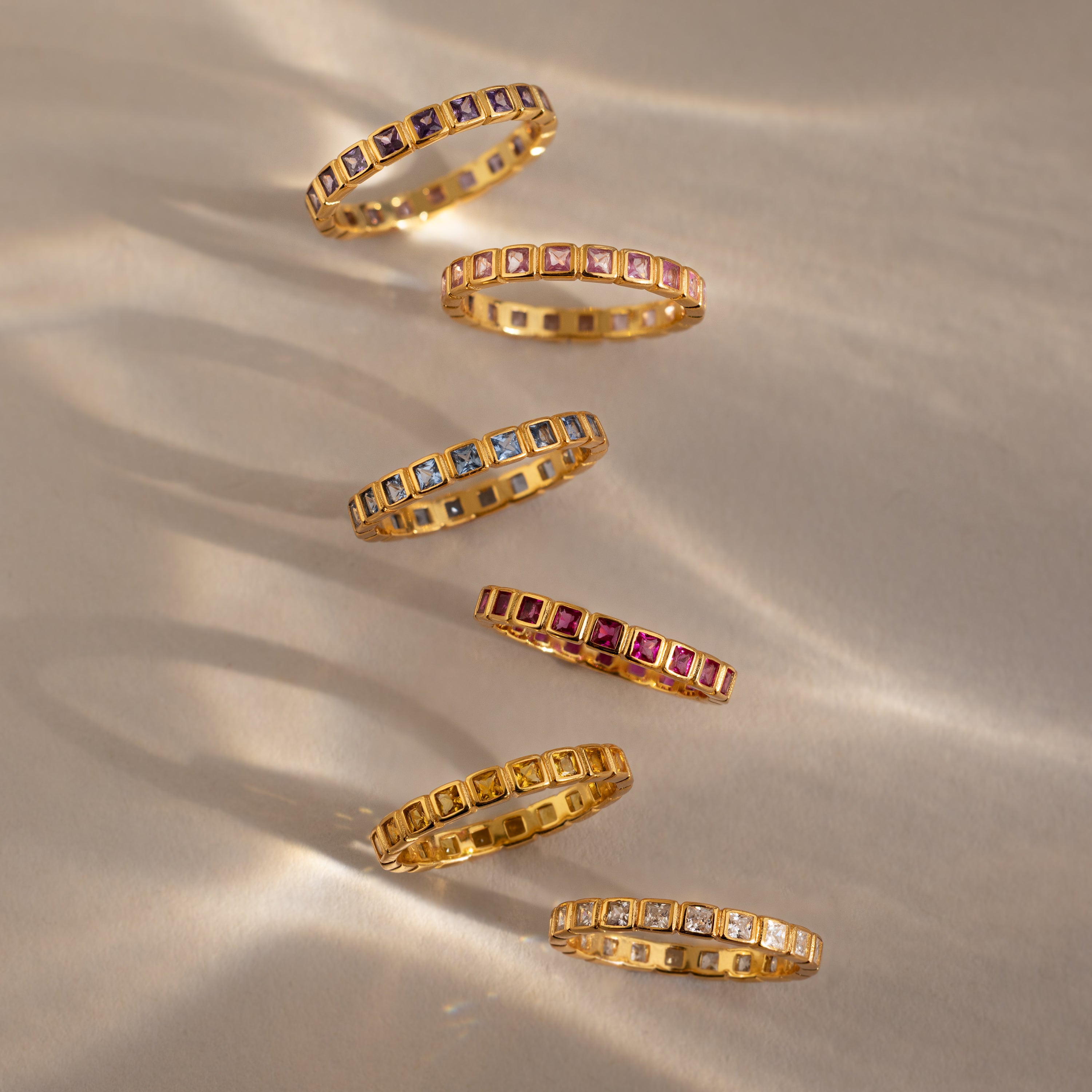 Six Eternity Bezel Peridot Rings with colorful square gemstones are arranged diagonally on a beige surface with soft shadows, highlighting the timeless elegance of this bezel birthstone ring.