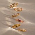 Six Eternity Bezel Birthstone Rings, each featuring a gold band and vibrant square-cut gemstones, are showcased on a neutral surface with soft shadows.