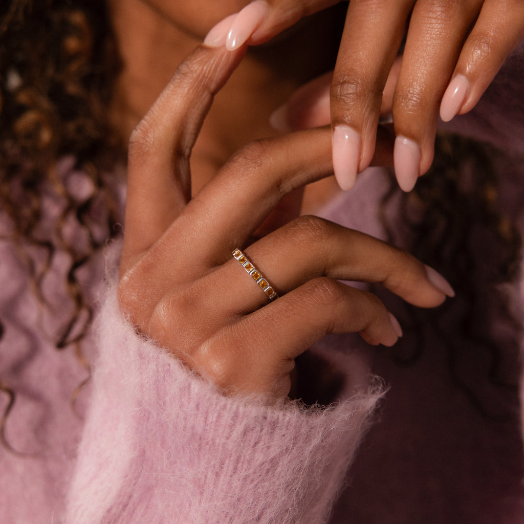 A hand with manicured nails wears an Eternity Bezel Aquamarine Ring, featuring square cut aquamarine gemstones, shown against a soft pink fuzzy sweater.