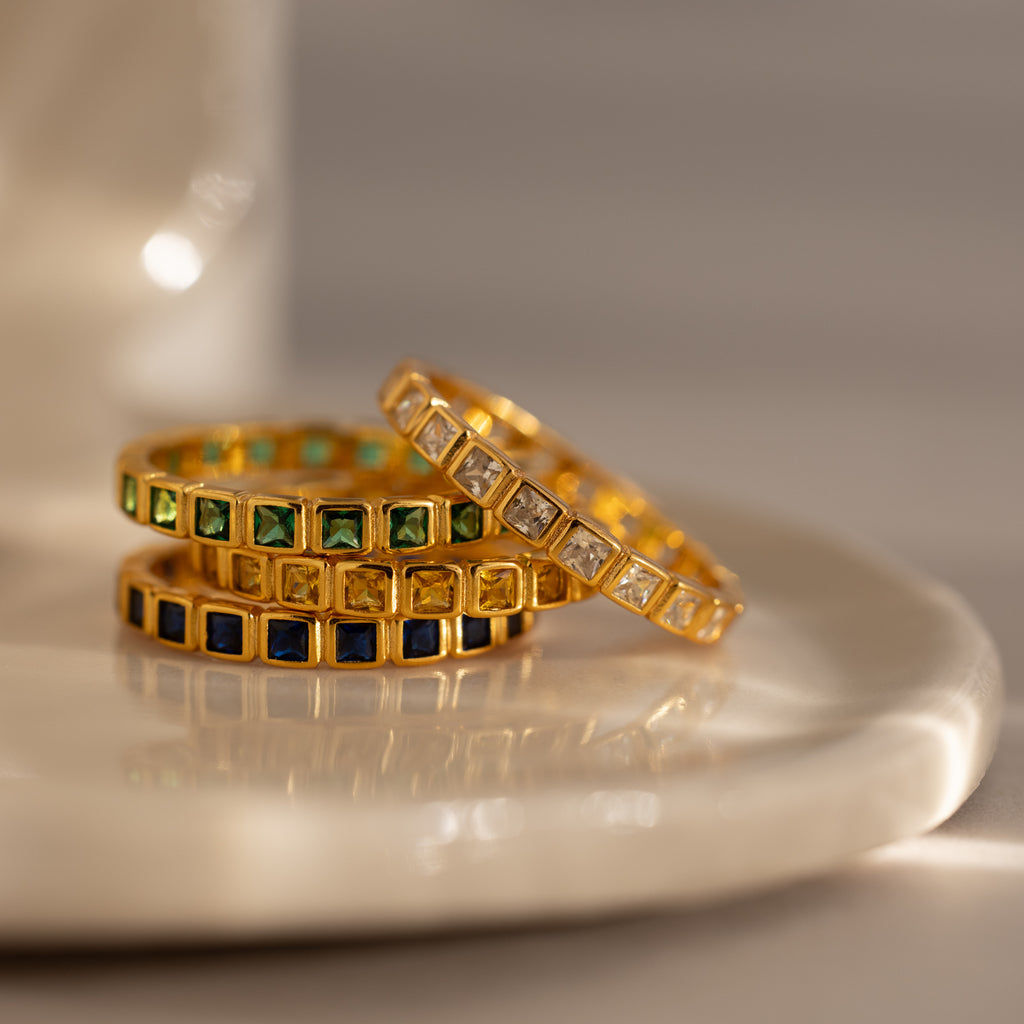 Three Eternity Bezel Sapphire Rings with square-cut green, blue, and clear gemstones are stacked on a glossy white surface.